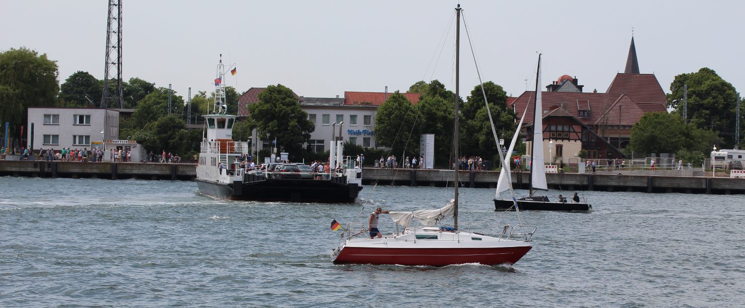 Foto: Autofähre legt ab, im Vordergrund segelt eine kleine Yacht vorbei