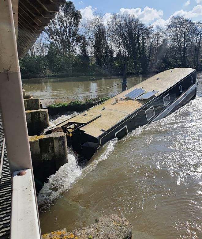Ein halb gesunkenes Hausboot ist gegen die Schutzpfähle eines Wehrs gedrückt, Wasser schäumt darum.