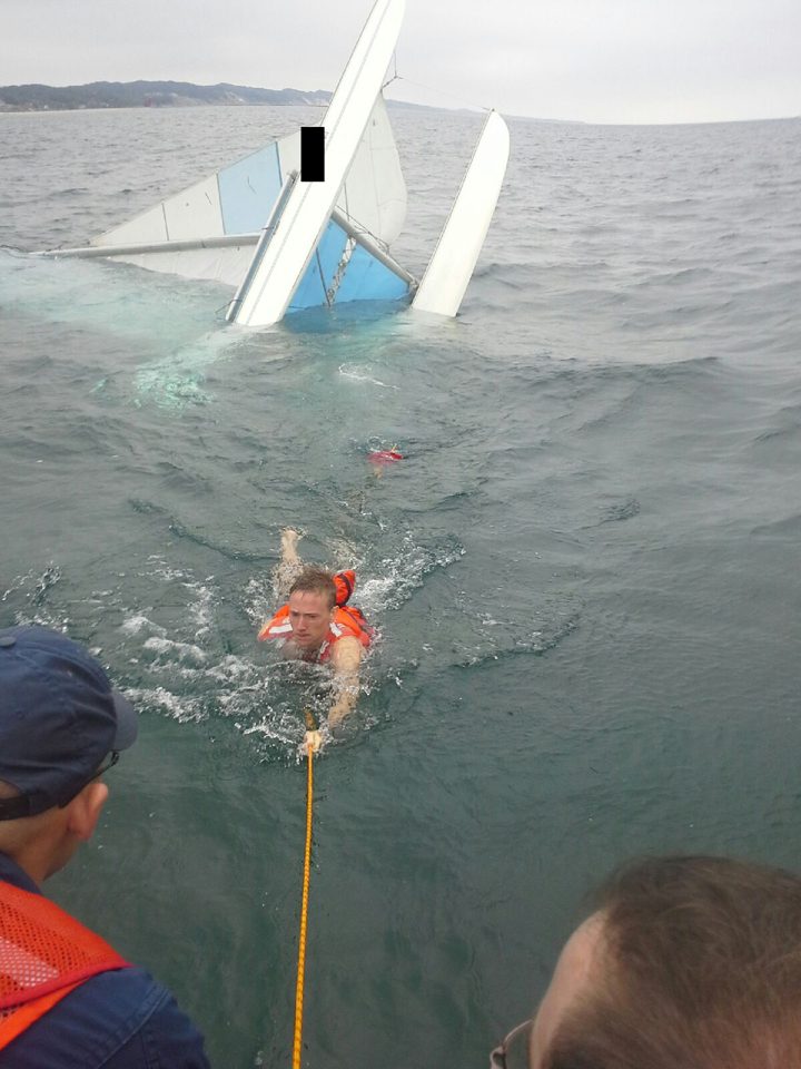 Ein Segler in orangefarbener Weste wird von einem Rettungsboot der Küstenwache aus dem Wasser geholt; im Hintergrund treibt das gekenterte Boot.